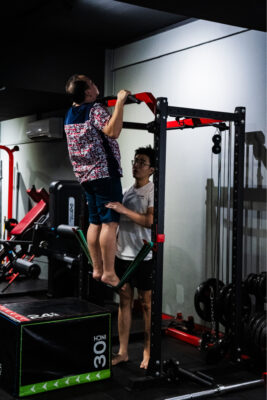 Senior male lifting himself on a gym pull-up bar with personal trainer supporting.