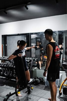 Woman performing exercises on dumbells with guidance from a personal trainer in a well-equipped gym in Singapore, focusing on strength and coordination.